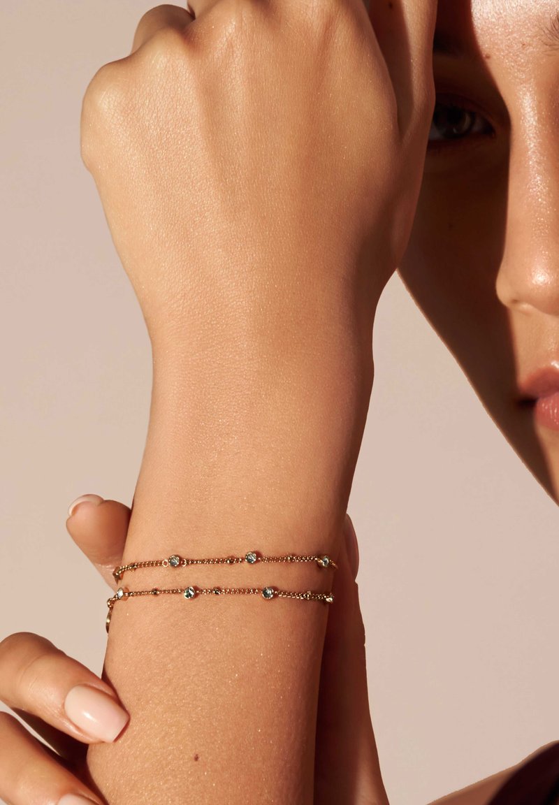 Bracciale d'oro con due catene, impreziosito da perline di turchese distribuite in modo uniforme, poggia su un braccio abbronzato. Il design è delicato e minimalista.