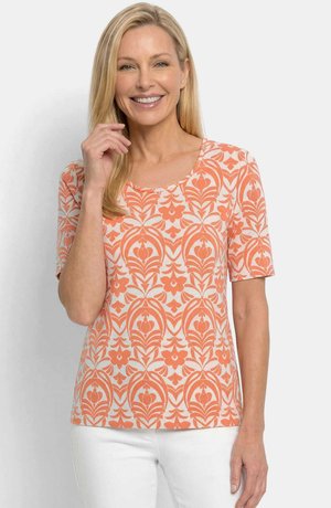 Lächelnde Frau mit langen blonden Haaren, die ein orange-weiß gemustertes kurzärmeliges Oberteil und weiße Hosen vor einem schlichten Hintergrund trägt.