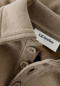 Polo de punto beige con una textura suave, tres botones con patrones florales y una etiqueta que dice "Lil'Atelier."