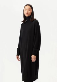 Svart poloklänning i mjukt tyg med långa ärmar och ribbade muddar, lös passform och knälång design. Minimalistisk stil.