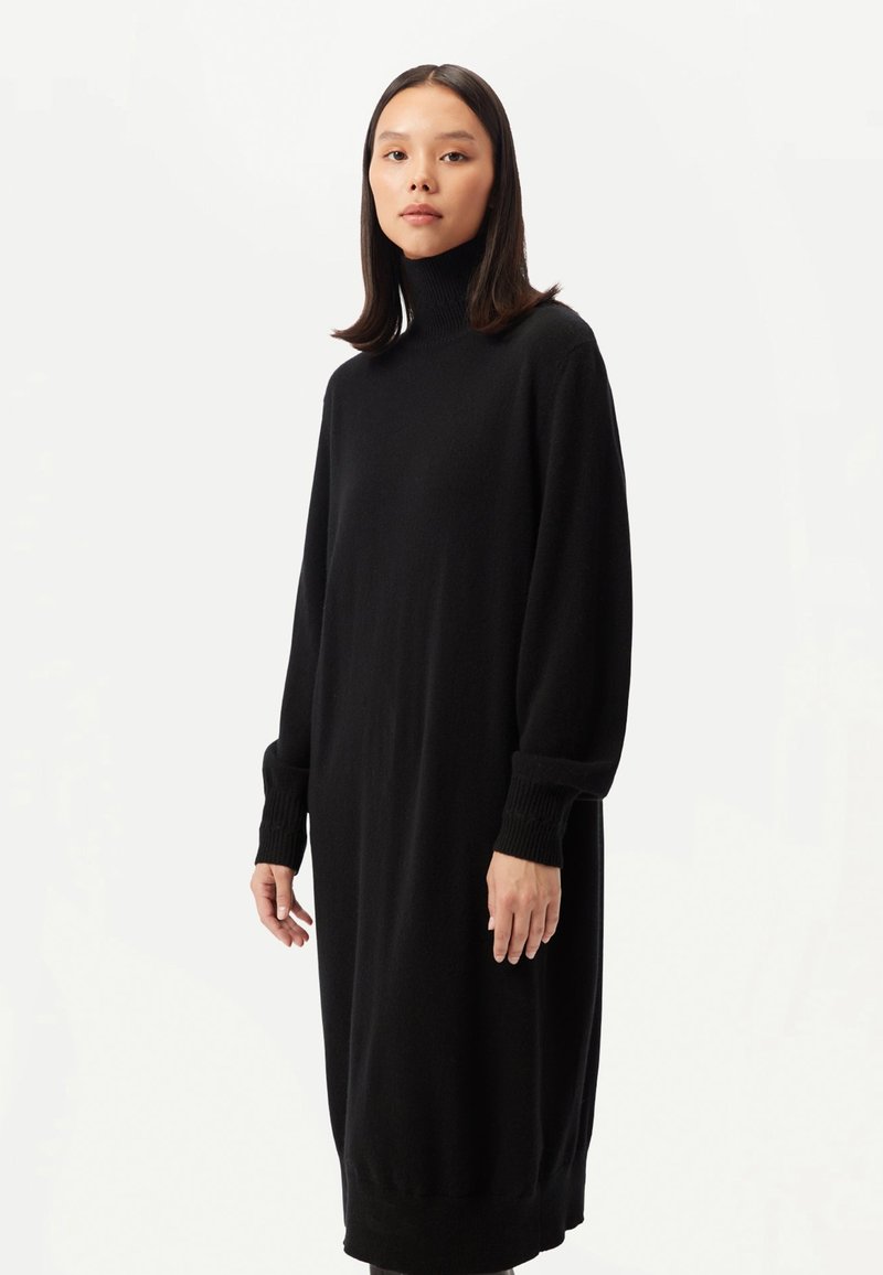 Svart poloklänning i mjukt tyg med långa ärmar och ribbade muddar, lös passform och knälång design. Minimalistisk stil.