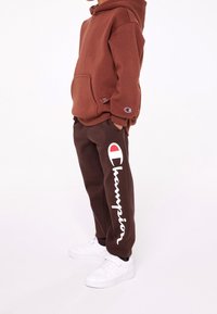 Felpa marrone con tasca frontale e toppa con logo, abbinata a pantaloni della tuta marroni con testo bianco "Champion". Sneakers bianche completano il look.