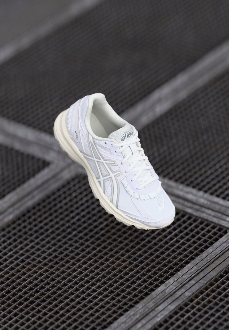 Chaussure de course blanche Asics avec lacets sur une surface en grille métallique sombre vue de dessus.