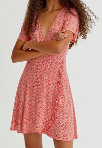 Femme les bras croisés portant une robe rouge à manches courtes avec un motif floral blanc et un décolleté en V profond.