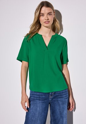 Blouse verte à manches courtes avec un décolleté en V, texture lisse et coupe décontractée, associée à un jean en denim bleu.