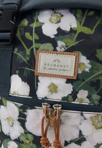 Gros plan d'un sac à dos à motif floral avec des fleurs blanches, des feuilles vertes, des tirettes de fermeture éclair marron et une étiquette cousue portant la mention "DOUGHNUT."