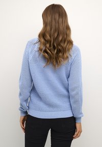 Una donna con capelli castani mossi indossa un maglione in crochet azzurro chiaro e pantaloni neri, in piedi di spalle contro uno sfondo semplice.