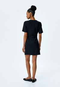 Zwarte jurk met korte mouwen, een aansluitende pasvorm en een riem rond de taille. De achterkant heeft een discrete ritssluiting. Zwarte platte schoenen maken de look compleet.