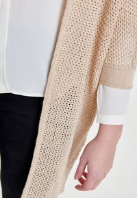 Světle béžový pletený kardigan s otevřenou vazbou a texturovaným povrchem, nošený přes jasně bílou blůzu s černým lemem.