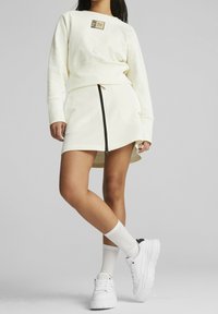 Felpa crema chiara con logo quadrato, abbinata a una gonna zip-front dello stesso colore. Sono indossate sneakers bianche chunky e calze bianche al ginocchio.