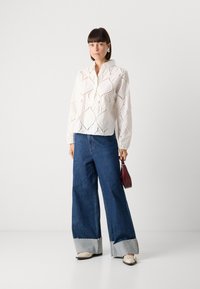 Blouse blanche brodée avec un motif en dentelle, assortie à un jean en denim bleu à jambes larges avec un ourlet effiloché et une finition en denim clair. Tenant un petit sac bordeaux.