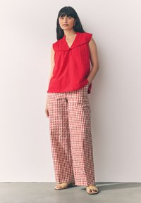 Rotes ärmelloses Oberteil mit gerafftem Kragen, kombiniert mit weit geschnittenen rot-weiß karierten Hosen im Gingham-Stil. Leicht strukturierter Stoff und flache Sandalen.