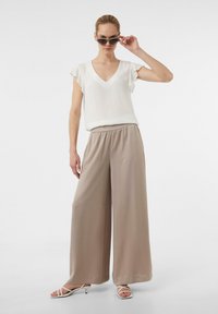 Witte v-hals top met korte gerimpelde mouwen, gecombineerd met lich beige wijde broek. Het model draagt grijze zonnebril en witte sandaaltjes met banden.