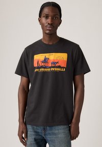 Mann i svarte Levi Strauss overaller og t-skjorte med oransje-gul western-inspirert grafikk, sammen med blå jeans, stående mot en enkel bakgrunn.
