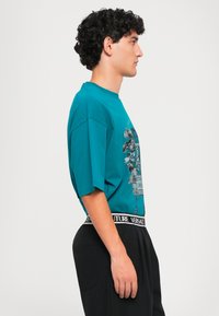 Fiatal férfi profilból, teális színű grafikás pólóban és fekete nadrágban, Versace Couture derékszárral, sima világos háttér előtt áll.