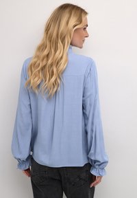 Light blue blouse featuring long, puffed sleeves with gathered cuffs, a relaxed fit, and a subtle texture. Back detail includes a yoke.