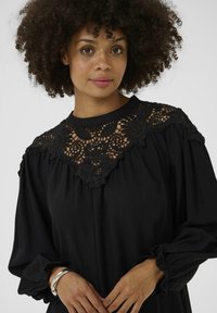 Blouse noire avec une encolure en dentelle ornée de motifs floraux complexes, manches longues avec poignets froncés et un col rond. Texture de tissu lisse.