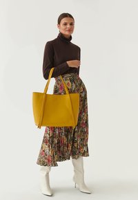 Bolso tote de cuero amarillo con dos asas largas, textura lisa, forma ligeramente curvada, al lado de una falda con estampado floral y botas de tacón blancas.