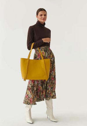 Borsa tote gialla in pelle con due manici lunghi, texture liscia, forma leggermente curvata, accanto a una gonna a fantasia floreale e stivaletti bianchi con tacco.