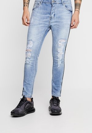 Vaaleansiniset pillifarkut, joissa on kulutettuja repeämiä, mustat sivuraidat ja nappisuljenta. Yhdistetty mustiin urheilukenkiin.