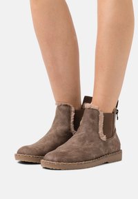 Bruna mockaankelstövlar med fårskinnskant och elastiska sidopaneler, utrustade med en gummisula och sydda detaljer för hållbarhet.