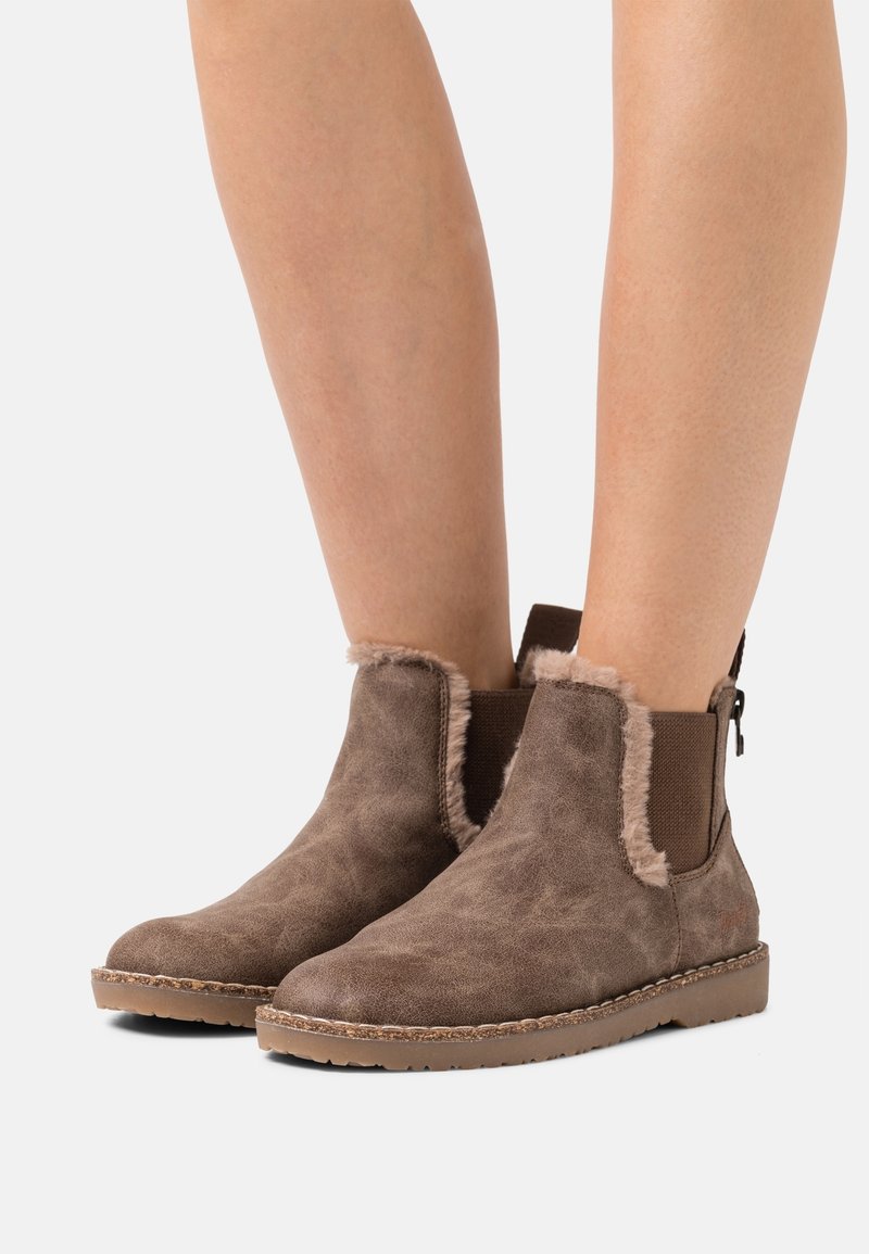 Bruna mockaankelstövlar med fårskinnskant och elastiska sidopaneler, utrustade med en gummisula och sydda detaljer för hållbarhet.