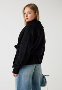 Sort cardigan med bred ribbet krave, bælte i taljen og lange ærmer, parret med lyseblå brede jeans. Glat tekstur, afslappet pasform.