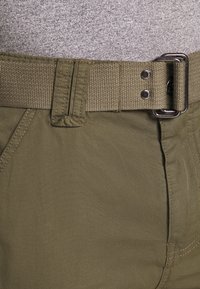 Olivegrüne Stoffhosen mit einem strukturierten, gewebten Gürtel. Zu den Merkmalen gehören ein klassischer Schnitt und sichtbare Nähte entlang der Seiten und des Bunds.