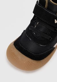 Scarpa nera in tela con tomaia in tessuto texture, cinturino in Velcro e suola in gomma marrone con righe orizzontali. Accentuata con dettagli del logo bianco.