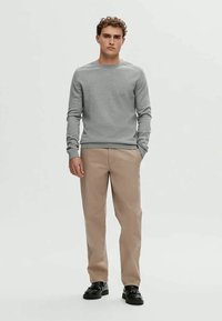 Hombre con cabello rizado que está de pie vistiendo una camisa de manga larga gris claro, pantalones beig y zapatos negros contra un fondo plano.