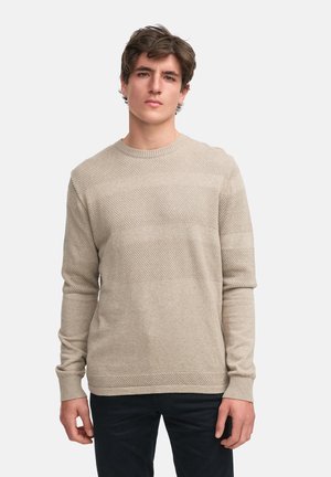 Beige strikkegenser med en teksturert mønster med horisontale striper, rund hals og lange ermer. Brukt med mørke bukser.