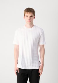 Tricou alb cu mânecă scurtă, cu un model texturat în diamant, guler rotund și croială dreaptă. Purtați cu pantaloni negri, expus pe un fundal simplu.