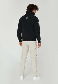 Svart bomberjacka med hög krage, ribbade mudd och nederkant, med logodetaljer bak och på ärmen. Modellen är ihopkopplad med ljusa byxor.