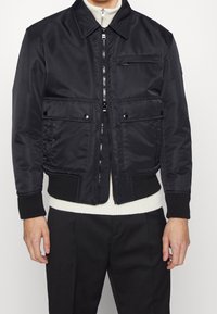 Svart bomberjacka med dragkedja fram, två bröstfickor och ribbade ärmslut. Tillverkad av ett mjukt tyg med hög krage och sidfickor.