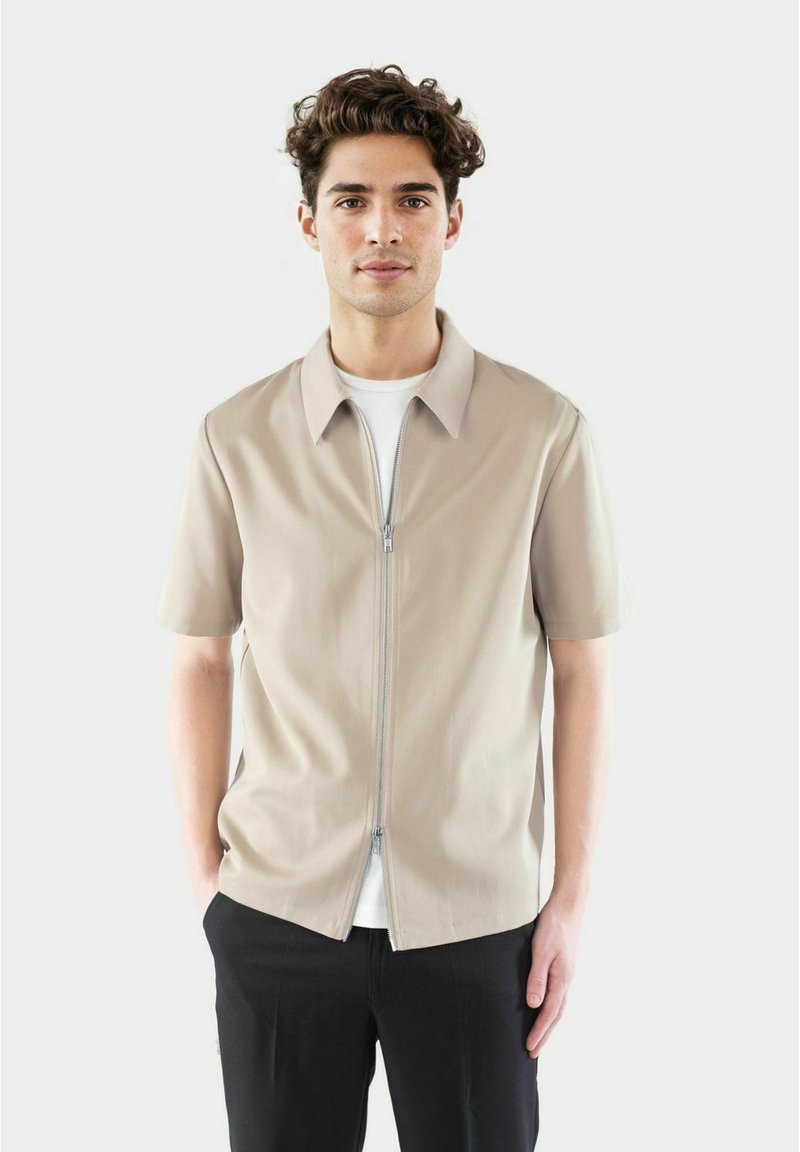 Kortermet beige zip-up skjorte med spiss krage, med en glatt tekstur og en avslappet passform, lagdelt over en hvit T-skjorte.
