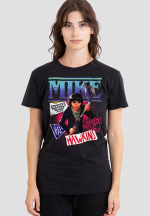 Young woman wears black T-shirt featuring "Mike" text, illustrated character holding a hockey stick, and "Stranger Things" and "Hawkins" graphics.