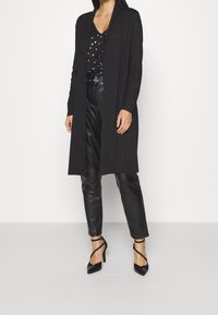 Lång svart cardigan i ett mjukt, texturerat tyg, matchad med en svart blus med prickar och svarta läderbyxor. Accentklackar med remmar.