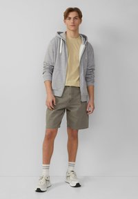 Jasnoszara bluza z kapturem na zamek założona na beżową koszulkę, w połączeniu z oliwkowymi szortami. Białe i szare sneakersy; pasiaki dopełniają stylizację.