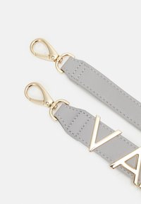 Poignée de sac en cuir gris avec des éléments en métal doré, présentant un détail de logo prominent et des bords cousus pour ajouter de la texture et de la durabilité.
