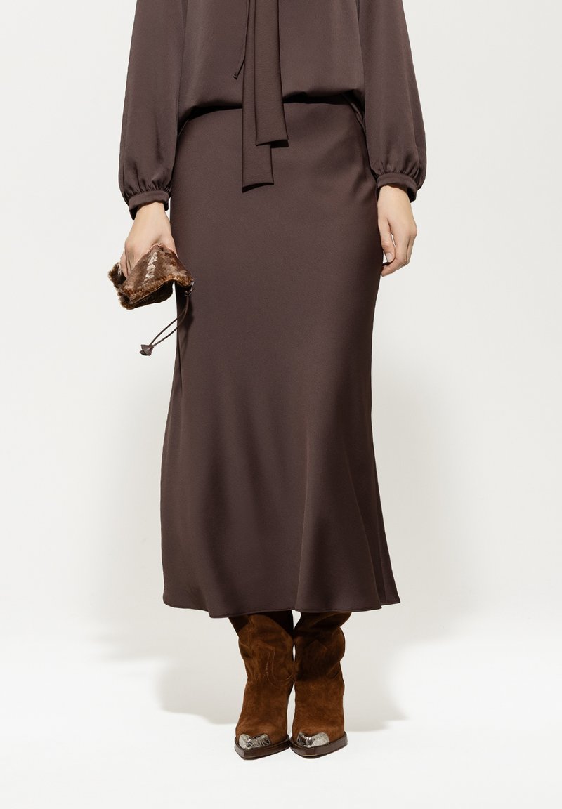 Brauer Satinrock mit schwingender Silhouette, kombiniert mit braunen Wildlederstiefeln bis zum Knie, die silberne Zehenakzente haben. Eine strukturierte braune Clutch wird in der Hand gehalten.