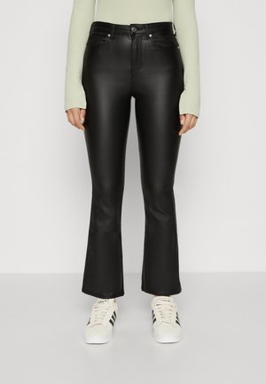 Person trägt schwarze Schlaghose aus Kunstleder, hellgrünes Langarmshirt und weiße Sneakers mit schwarzen Adidas-Streifen.