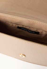 Μπεζ πορτοφόλι με υφή, ανοιχτό για να δείξει το λείο ύφασμα στο εσωτερικό του με μαύρη ετικέτα που γράφει "ANNA FIELD" και έναν χρυσό μαγνητικό κούμπωμα στη φλάντζα.