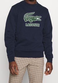 Námořnická mikina s zeleným krokodýlím logem a nápisem "LACOSTE" tučnými písmeny. Má žebrované manžety a lem, vyrobena z měkkého bavlněného materiálu.