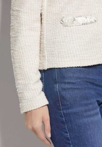 Texturiertes Cremepullover mit Rundhalsausschnitt, ausgestattet mit einem Paillettenakzent auf der linken Tasche, kombiniert mit engen blauen Jeans.