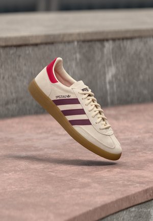 Μπεζ αθλητικό παπούτσι Adidas Spezial με μπορντό ρίγες, κόκκινη φτέρνα και καουτσουκένια σόλα, εμφανιζόμενο στον αέρα πάνω από πετρώδη επιφάνεια.