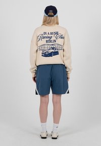 Person trägt einen beigen Pullover mit der Aufschrift "Racing Club Berlin" und einem Auto-Grafik, blaue Shorts, weiße Socken, weiße Schuhe und eine blaue Kappe, Blick nach hinten.