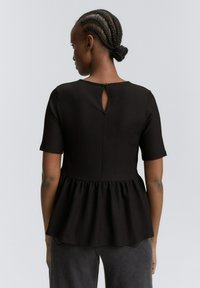 Top peplum noir à manches courtes, en tissu texturé, avec un détail de dos en forme de clé et un bas évasé, associé à un pantalon gris.