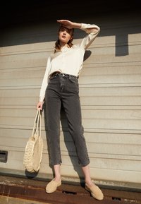 Blouse crème avec des boutons, jeans taille haute gris, espadrilles beiges et sac en osier texturé. Le mannequin pose devant un mur texturé.