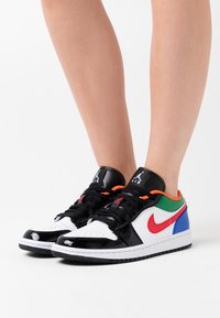 Zapatillas en blanco y negro con detalles de cuero brillante, paneles laterales multicolores (rojo, verde, azul, naranja) y cordones negros.