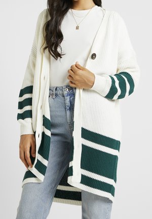 Vrouw draagt een witte geribbelde lange cardigan met groene en witte strepen, een wit topje, lichtblauwe spijkerbroek en een gouden hangerketting.
